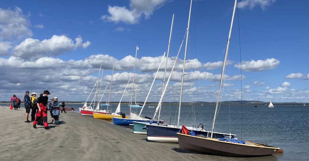 A row of dinghies pulled up on a sandy beach in full sun with bubbly clouds.